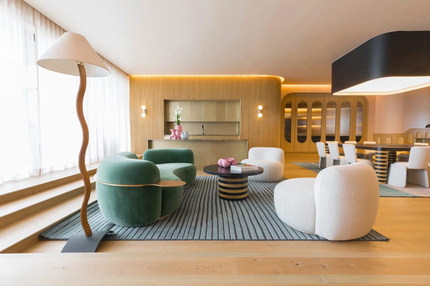 Modern living space with a green sofa, white chairs, and a striped rug. A wavy floor lamp stands by large windows, creating a warm, inviting ambiance. By one of the best mexican interior designers.