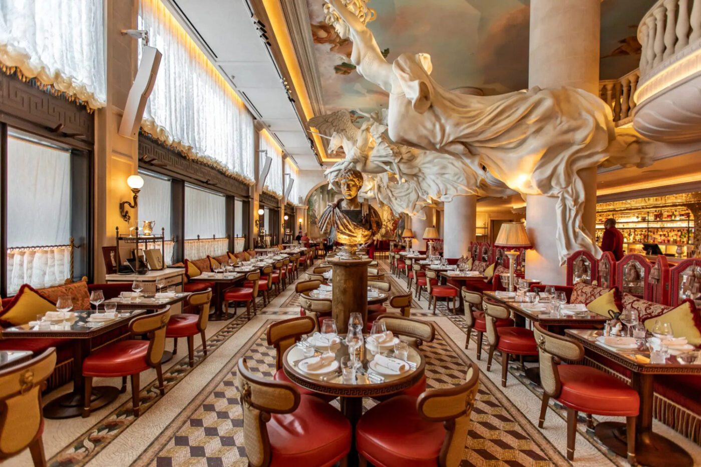 Bacchanalia-Restaurant-London-Mayfair-Interiors Elegant restaurant interior with ornate decor featuring a large white sculptural installation and classical bust. Rows of tables with red chairs create a luxurious ambiance. One of the best designed restaurants in London