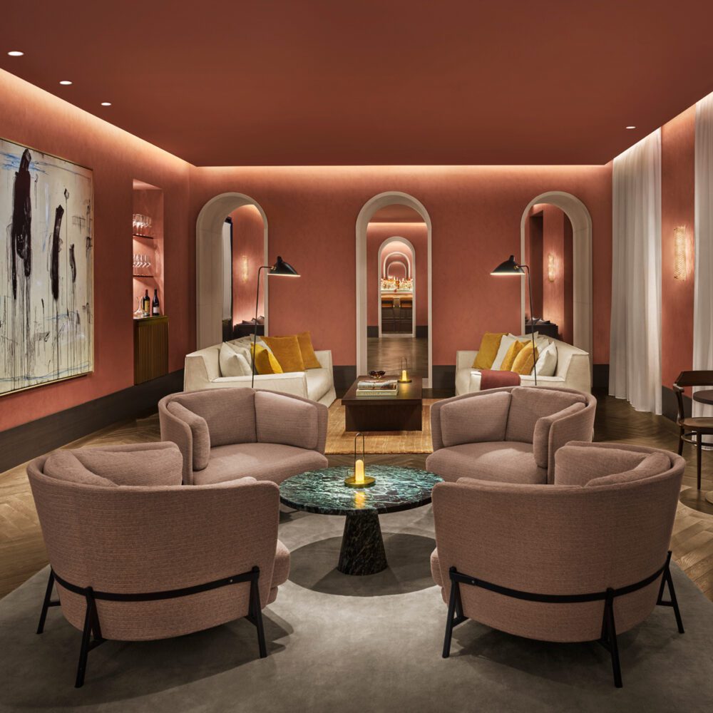 Como1_17_RGB_V4-1 by edition hotels Hotel lounge with peach walls and archways. Features include plush armchairs, a marble table, abstract art, and warm lighting, creating a cosy ambiance.