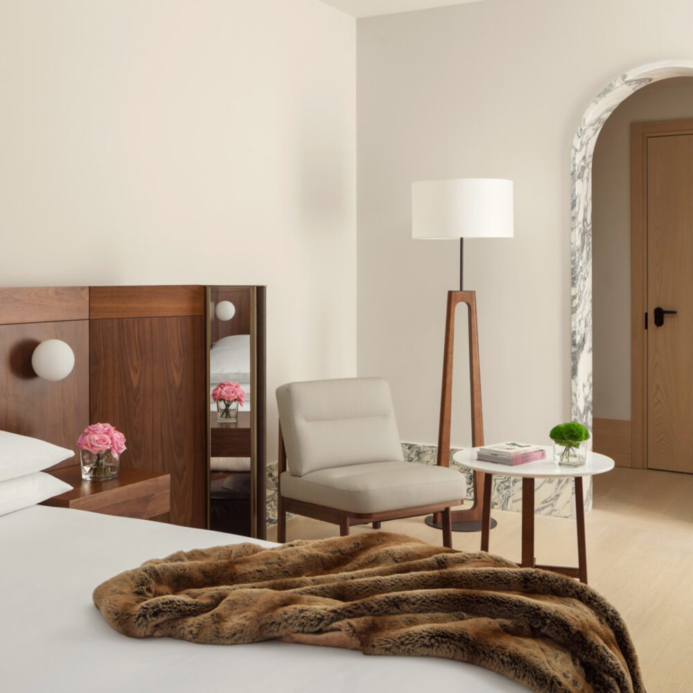 EDT-MILLE-Guest_Room_02 by edition hotels A hotel bedroom with a bed, wooden headboard, and white pillows. A cosy brown throw lies on the bed. Beside it, a pink flower vase sits on a nightstand. Nearby, a beige chair and a white round table with a green plant add elegance. A tall wooden floor lamp stands next to a marble archway, creating a serene and stylish atmosphere.