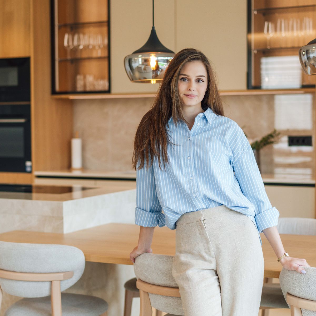 Pia Podlipnik, Estilia Design interior designer dressed in a blue shirt and white pants, stands in a stylish kitchen, surrounded by modern appliances and decor.