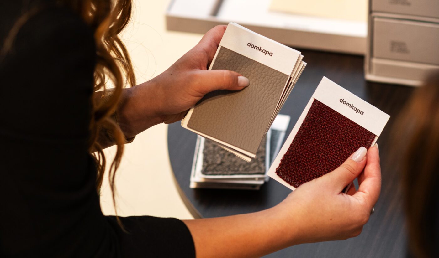 Fabrics by Domkapa Sample Box A woman holds up a pair of fabric swatches, showcasing their vivid designs and playful colour and texture combinations. About furniture colours and fabrics