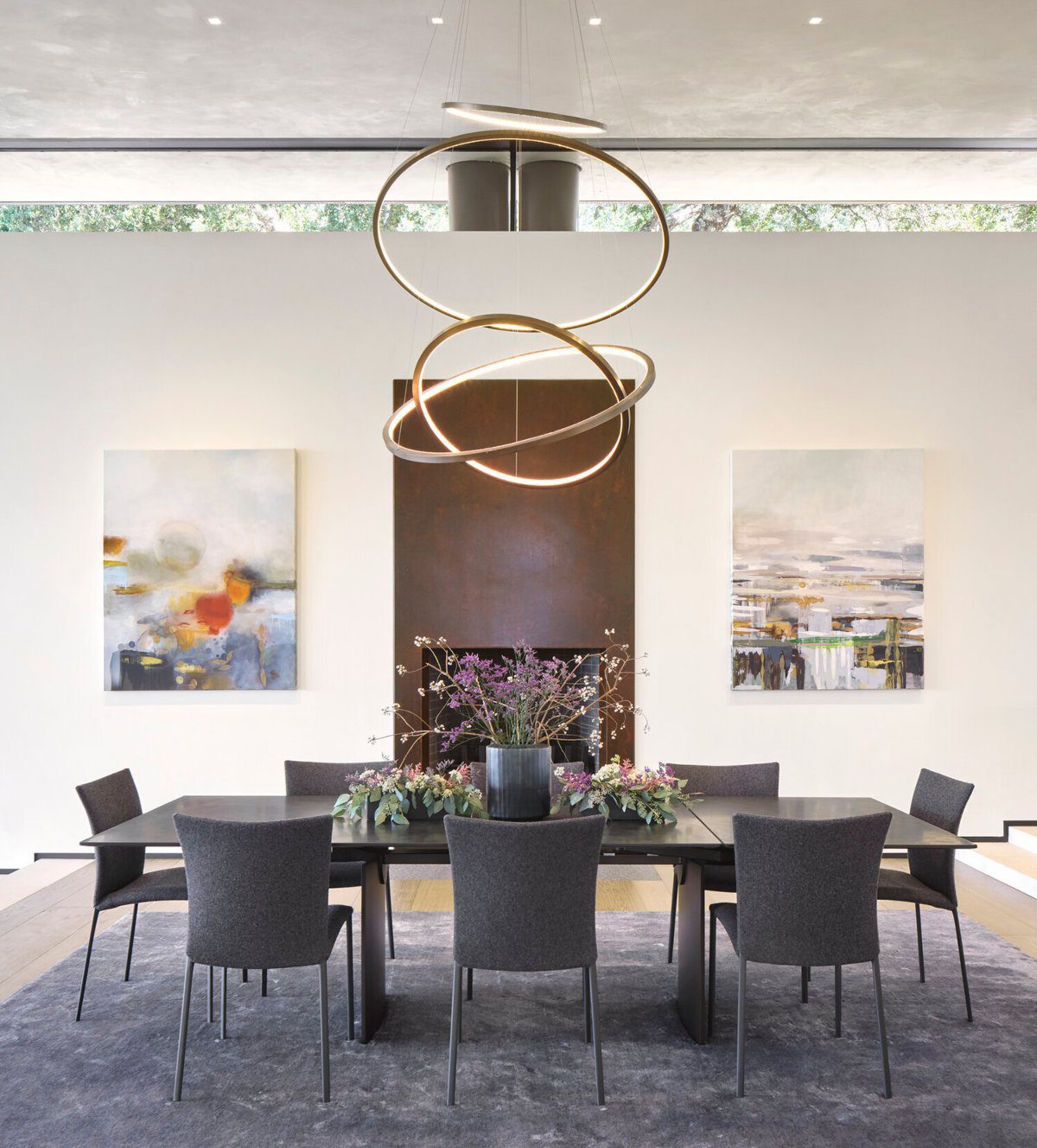 Forest Aerie Project by Shawback Design Forest Aerie Project by Shawback Design. The dining room is in white and grey tones with some gold accents in the lighting. It has a wood dining table with six dining chairs around it. In the middle of the dining table it has a vase with flowers.