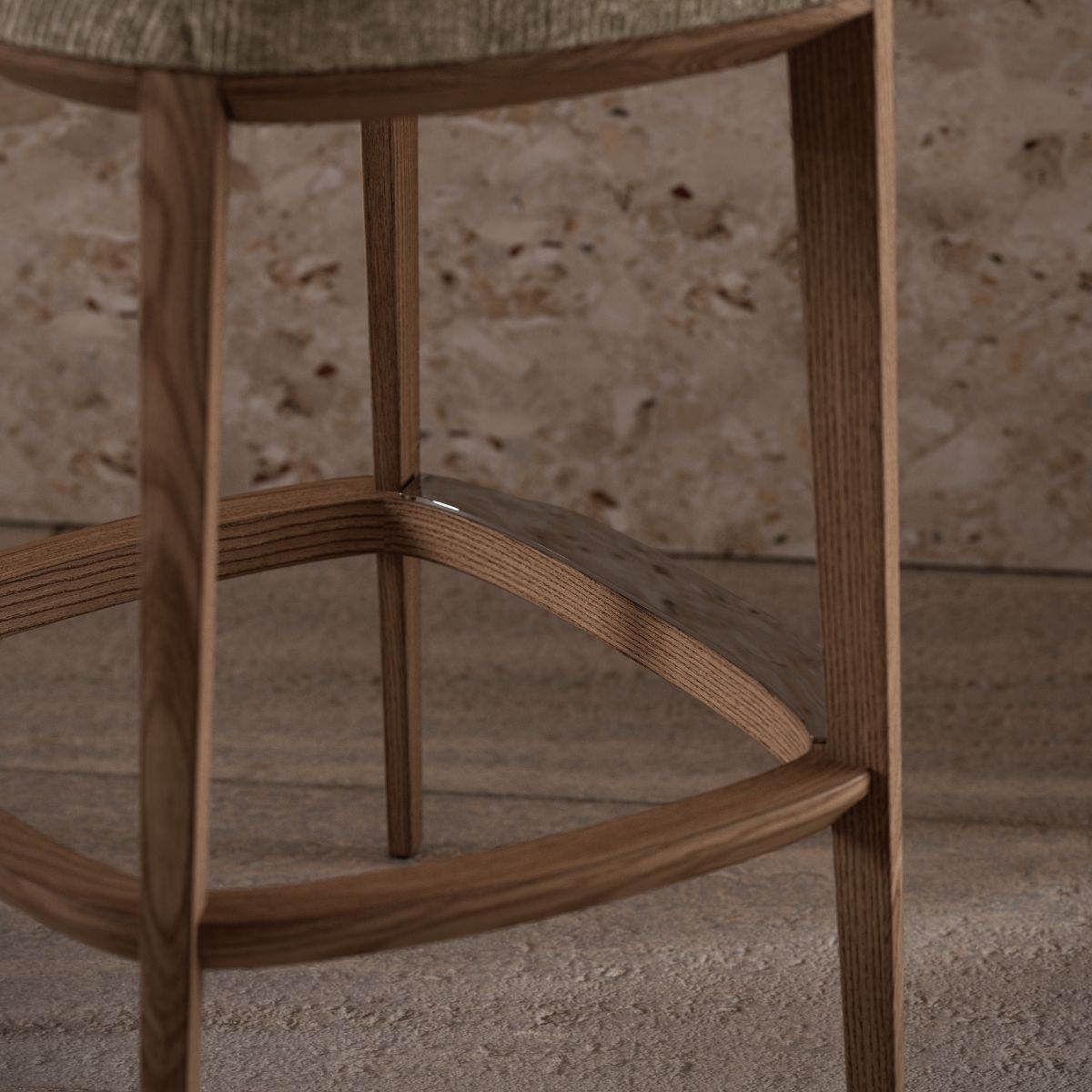Jermain Bar Chair by Domkapa (4) Close-up of a wooden stool leg and footrest, set against a textured stone background. The natural wood tones evoke a warm, rustic feel.