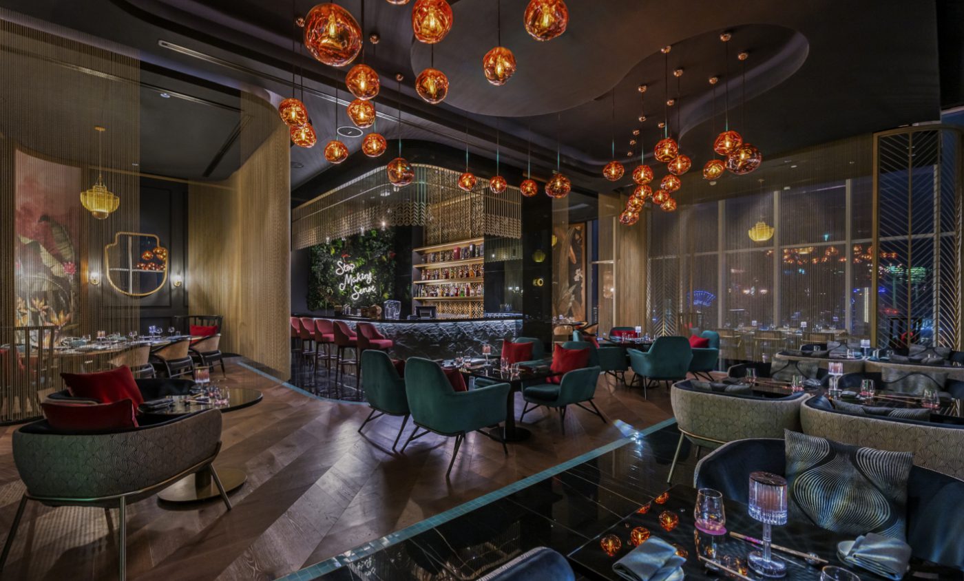 Keane Brands Restaurant design in dark tones. The chairs are in dark green and others with a floral pattern. The lights are orange and suspended in the ceiling. With a bar in the corner with red bar chairs.