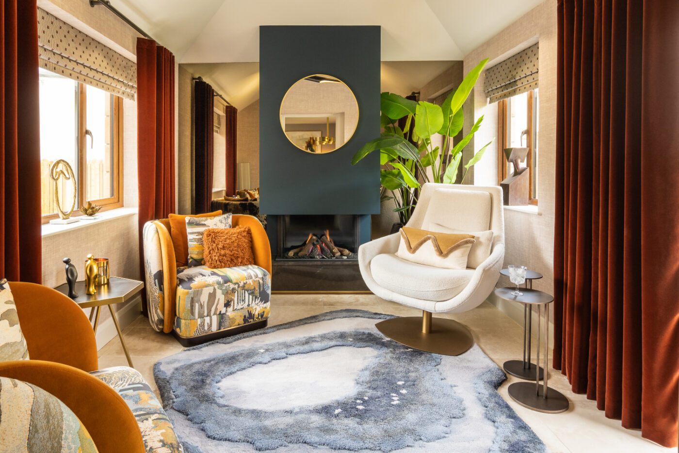 Kris Turnbull Project - Peter Bruce Photography A cosy living room with a modern fireplace, round mirror above it, and large green plant beside it. The space includes a white swivel chair, patterned armchairs, and a textured rug in shades of blue and gray, with large windows draped in burgundy curtains.