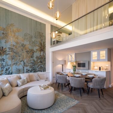 La Fenice Interiors project Living room and dining room with a white sofa, white pouf, oval dining table and six brown dining chairs. With a wall with a blue print and gold accents.