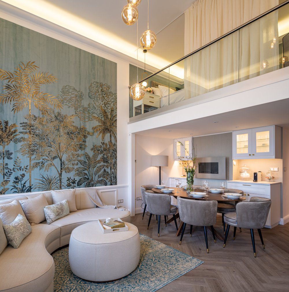 La Fenice Interiors project Living room and dining room with a white sofa, white pouf, oval dining table and six brown dining chairs. With a wall with a blue print and gold accents.