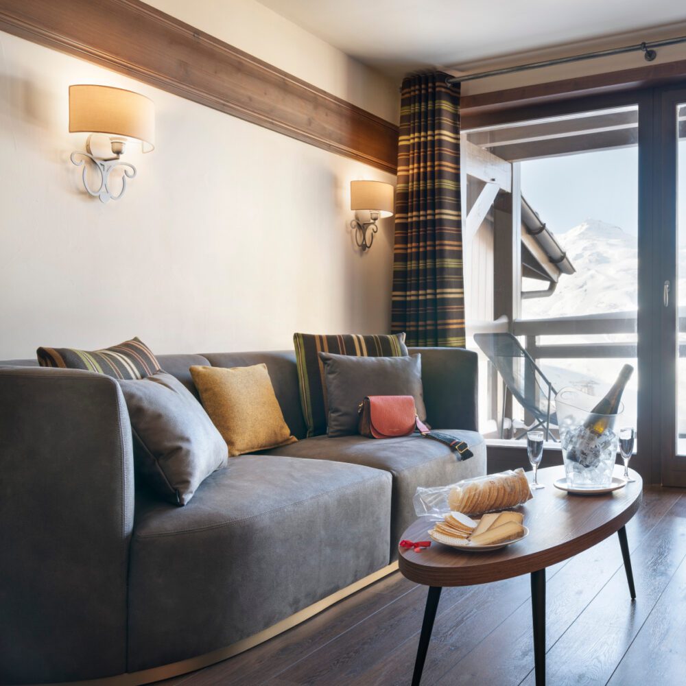 Le Hameau du Kashmir Suite with Chloe Bright living room scene showcasing a soft sofa, a chic table and a large window inviting sunlight inside. From Le Hameau du Kashmir hotel in the french alps.