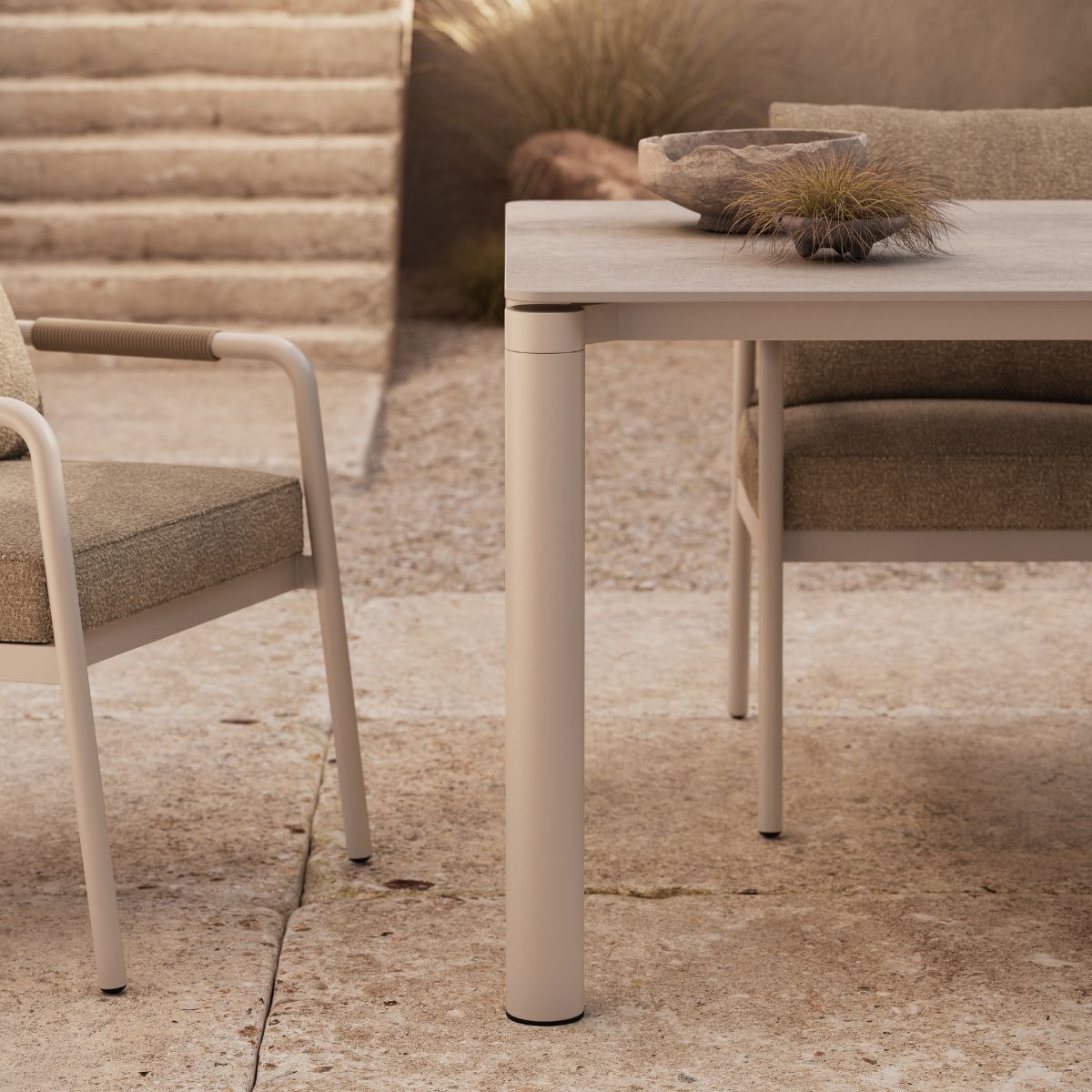 Pontus Dining Table by Domkapa (3) Outdoor patio scene with a beige table and chairs on a stone surface. A decorative bowl and grass are on the table. Steps and plants are in the background.