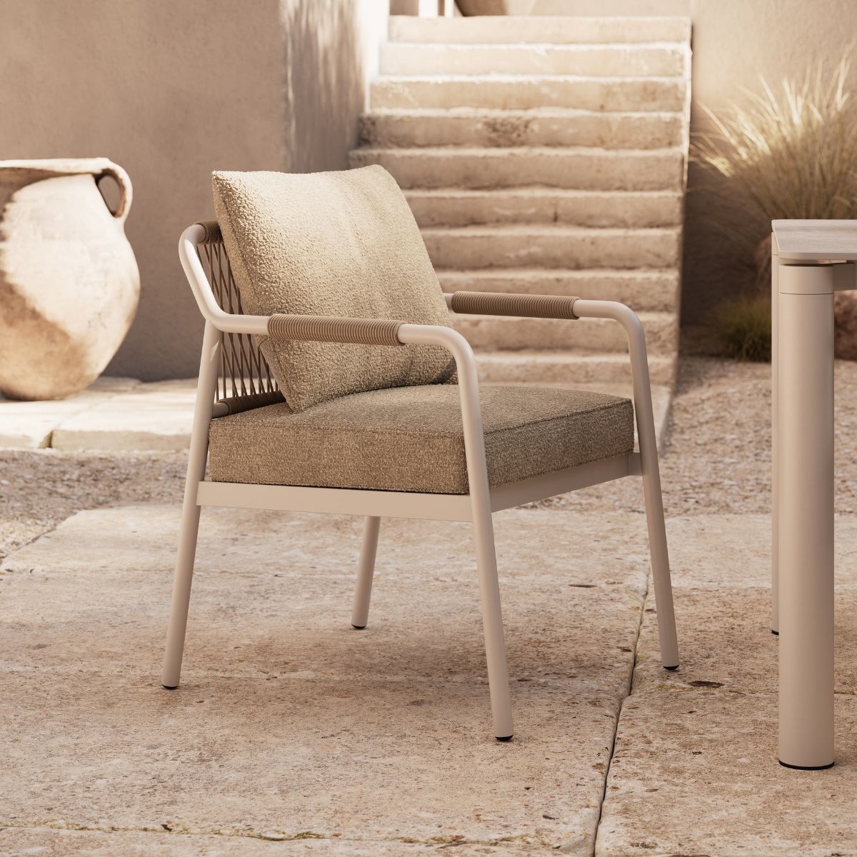 Neroli Dining Chair by Domkapa A stylish beige dinng chair with padded cushions sits on a stone patio near rustic stairs. An amphora stands nearby, creating a serene, Mediterranean vibe.