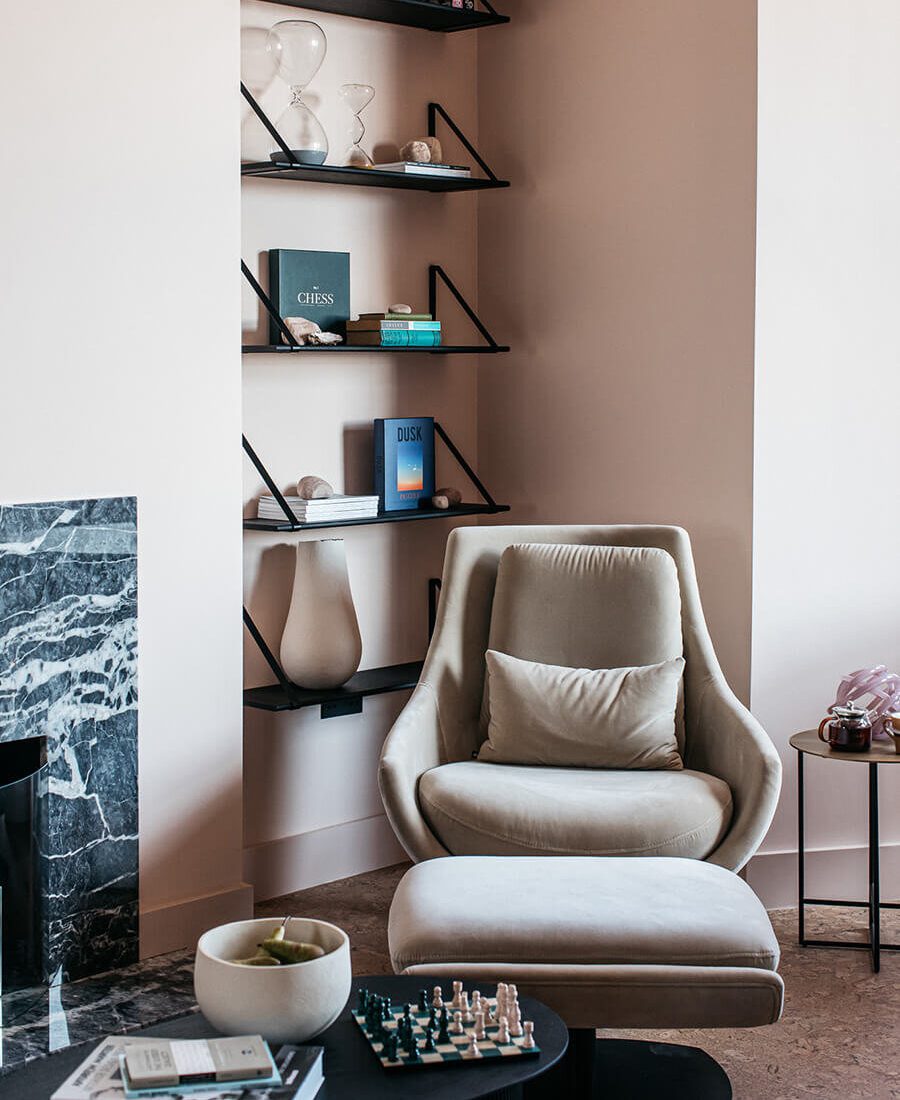 Port Hotel Design A minimalist living room with soft pink walls features a modern beige sofa, round chairs, and a cosy fireplace. Shelves display decorative items, and a dining area with a table and chairs is visible through an archway. Port Hotel