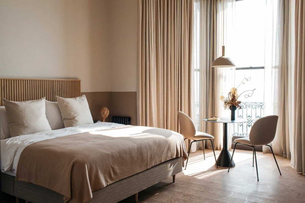 Port Hotel Double Room A cosy, sunlit bedroom features a large bed with beige linens and white pillows. To the side, a small round table with two chairs sits by a window with soft curtains. A contemporary lamp hangs above the table, enhancing the inviting atmosphere.