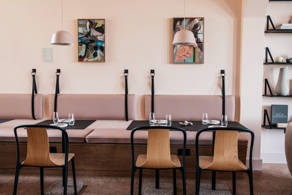 Port Hotel Restaurant A minimalist living room with soft pink walls features a modern beige sofa, round chairs, and a cosy fireplace. Shelves display decorative items, and a dining area with a table and chairs is visible through an archway.