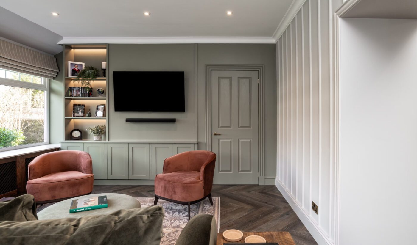 Project-by-Zhanar-Design-Photography-by-Niall-Hastie-Photography A cosy living room with pastel green walls, a wall-mounted TV, two plush pink chairs, and a built-in shelf with books and decor. The atmosphere is inviting.