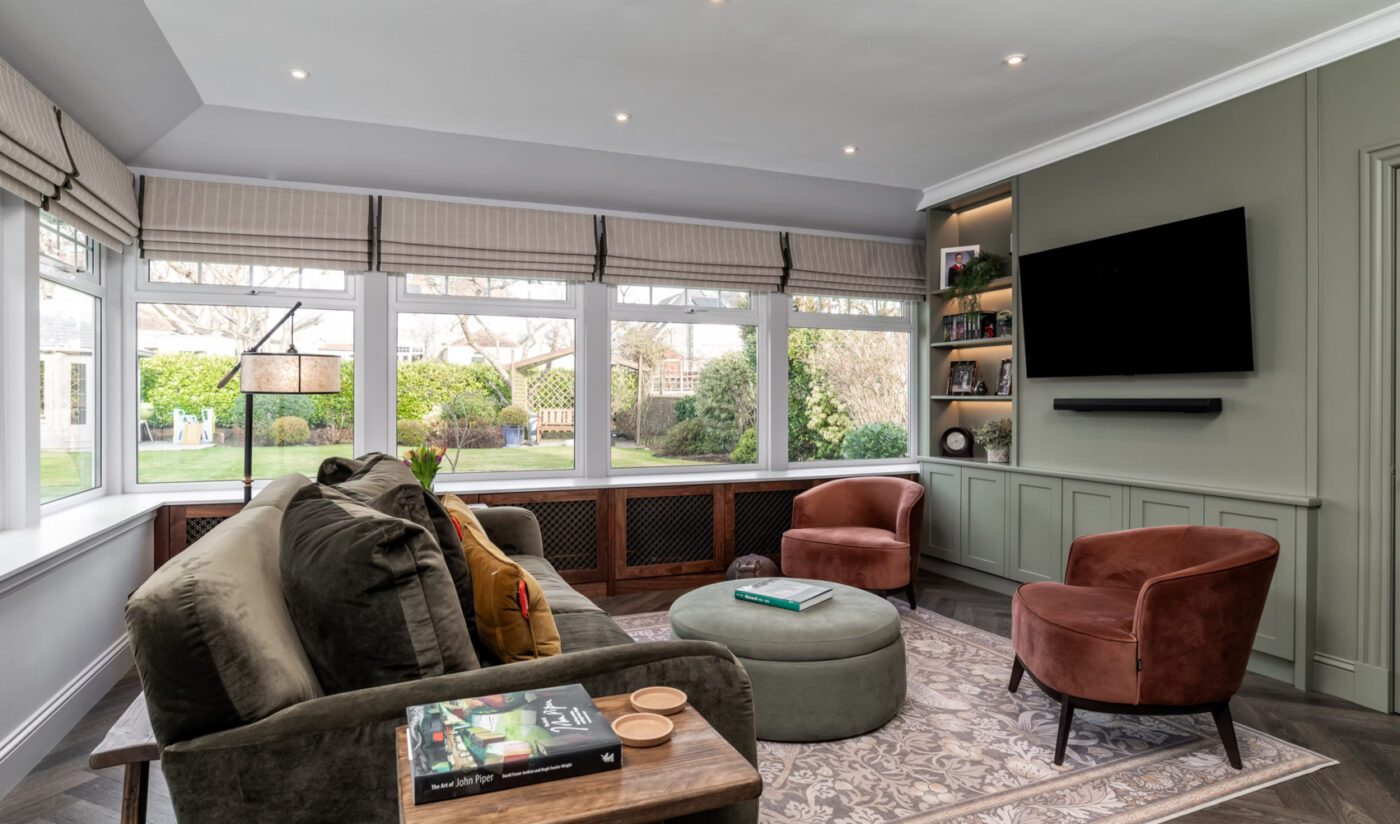 Project by Zhanar Design - Photography by Niall Hastie Photography Spacious living room with large windows overlooking a garden. Features green sofa, red armchairs, pouf, wall-mounted TV, and bookshelves. Cosy ambience. Project by Zhanar Design.