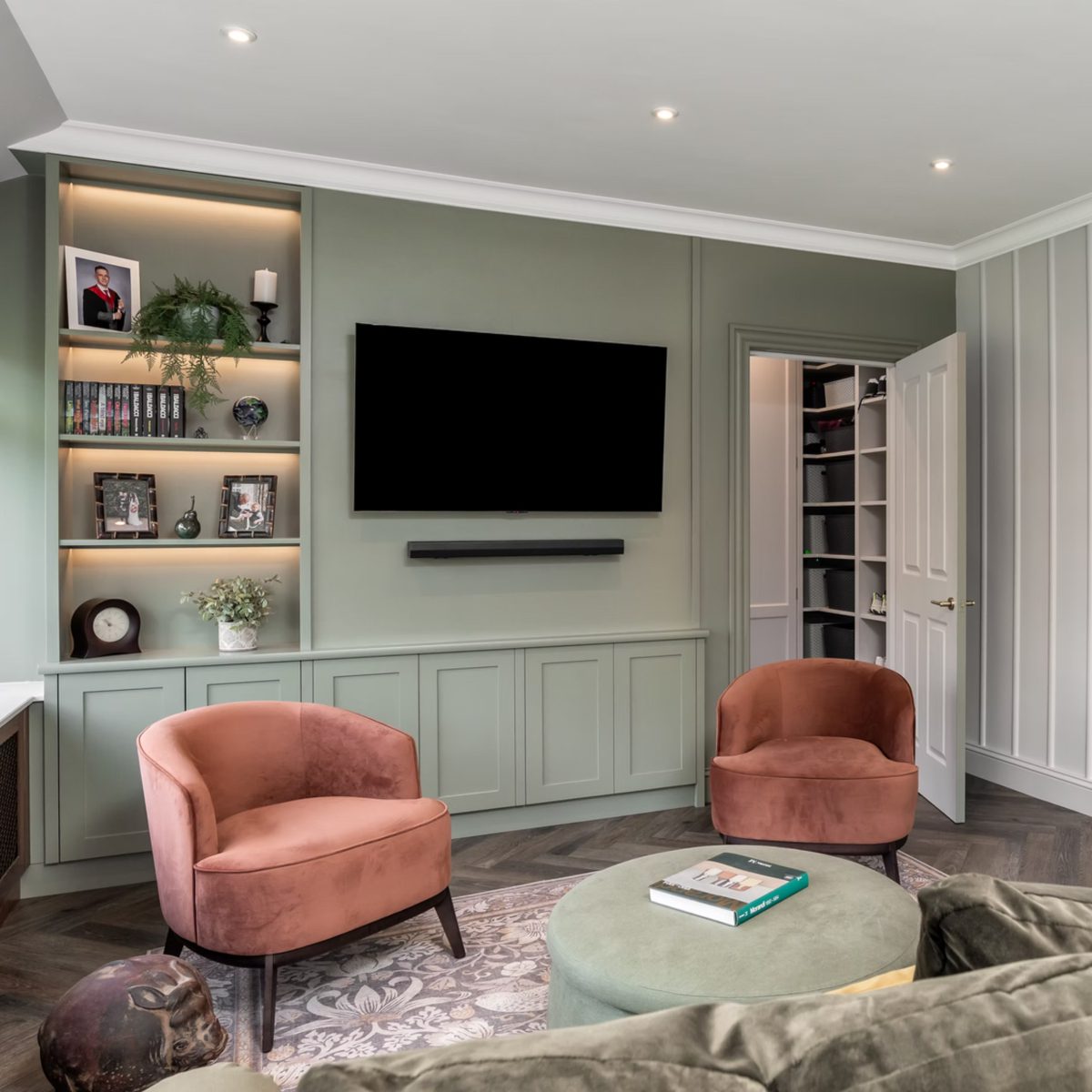 Project-by-Zhanar-Design-Photography-by-Niall-Hastie-Photography-3 Modern living room with muted green walls, two salmon pink armchairs, a wall-mounted TV, open door revealing shelves, and decorative bookshelves. Cosy ambiance. Project Designed by Zhanar Design.