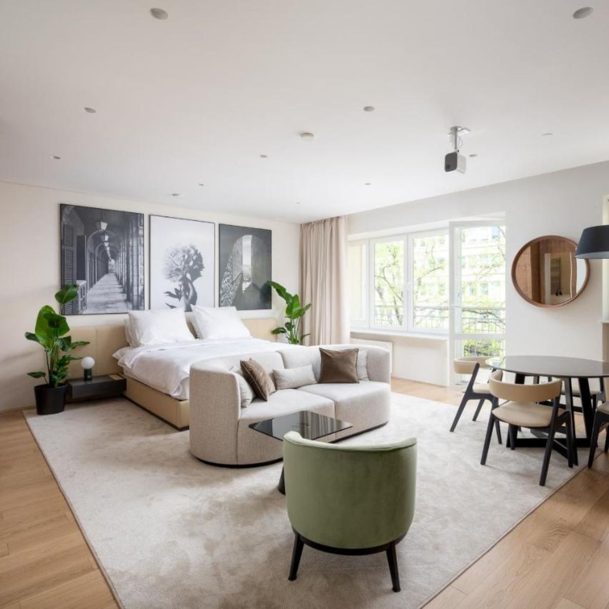 A modern, airy hotel suite features a bed with white linens, abstract art, and indoor plants. A cozy seating area and dining table create a serene, inviting space.