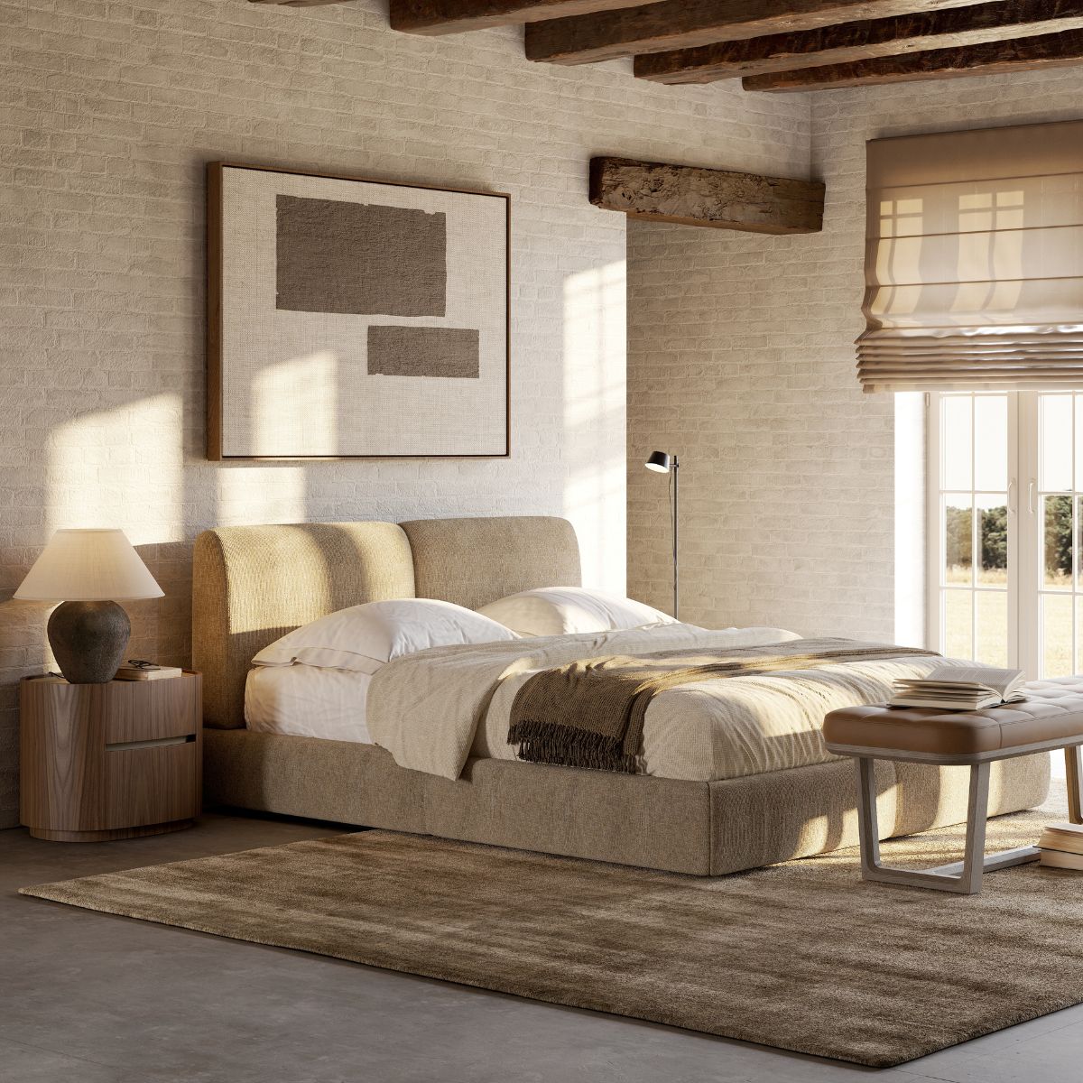 A modern bedroom with a beige upholstered bed, wooden nightstand and lamp, large window with Roman shade, bench and abstract art on a brick wall.