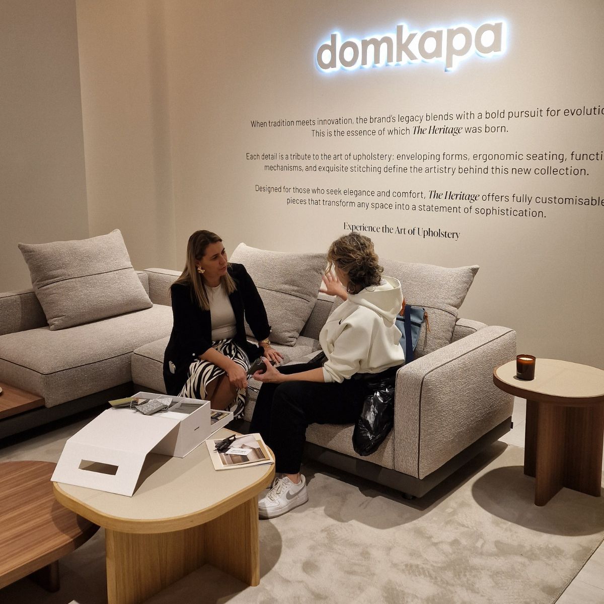 choose the perfect Domkapa sofa (3) Two women sit on a modern grey sofa in a stylish showroom, engaged in conversation. A wall with branding and text about upholstery is behind them.