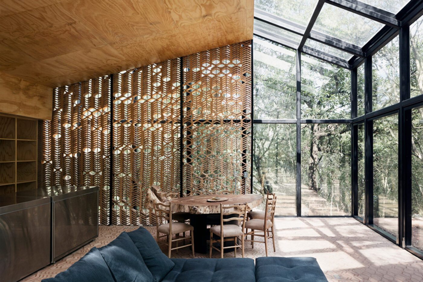 Stylish modern interior with a blue sofa, wooden lattice wall, and glass ceiling overlooking trees. The space feels airy and connected to nature.