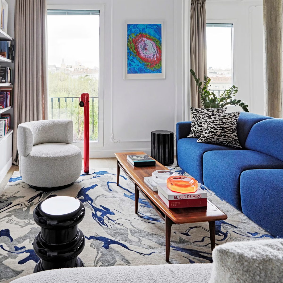 imgi_171_salon-sofa-azul-68d389c6d535c Modern living room with a bright blue sofa, a patterned black-and-white pillow, and a round white chair. A colourful abstract painting hangs between two large windows, and a wooden coffee table displays books and an orange glass object. The space is vibrant and contemporary. Project by Blanca Rosa Gutiérrez Studio