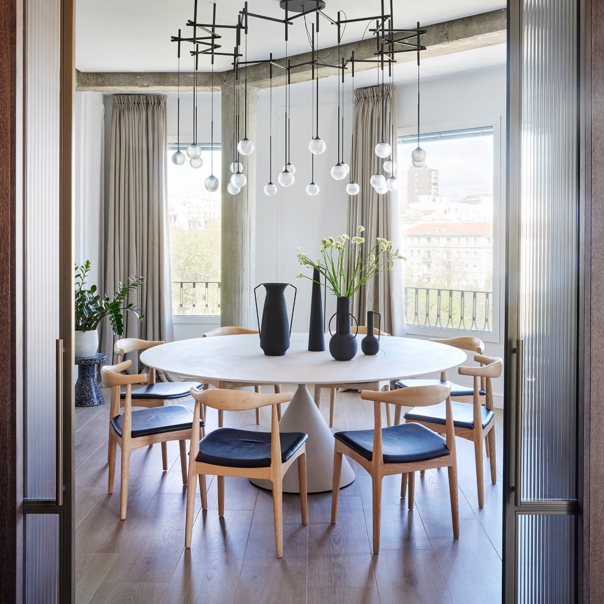 imgi_173_comedor-68d389c1c3a6b Modern dining room with a round white table, surrounded by wooden chairs with navy cushions. Black vases and flowers are on the table. Large windows and a geometric chandelier create an airy, stylish ambiance. Project by Blanca Rosa Gutiérrez