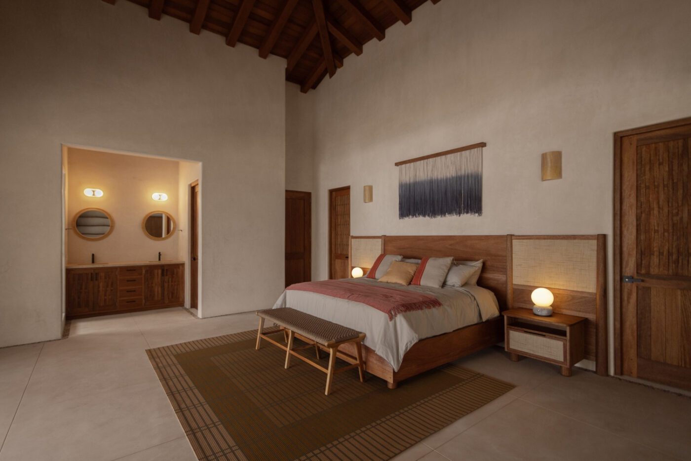 Cosy bedroom with high ceiling, wooden bed and earthy tones. Soft lighting, two doors, a woven wall hanging and a glimpse of a bathroom are visible. By one of the best mexican interior designers.