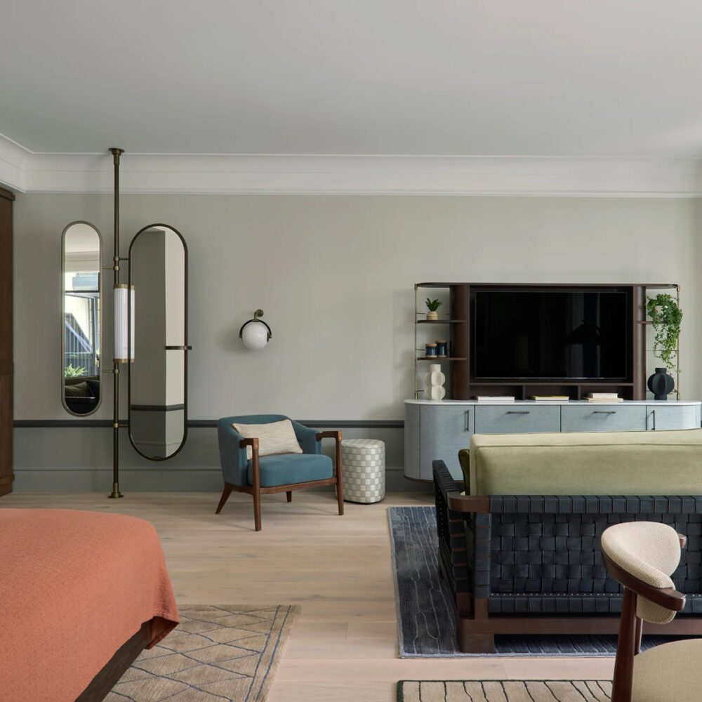 imgi_71_Courtyard_Terrace_Suite_Living_Room by six senses Stylish modern living room in a hotel with neutral tones. Features a blue armchair, woven-back sofa, wall-mounted TV, mirrors, and elegant decor, creating a calm ambiance.