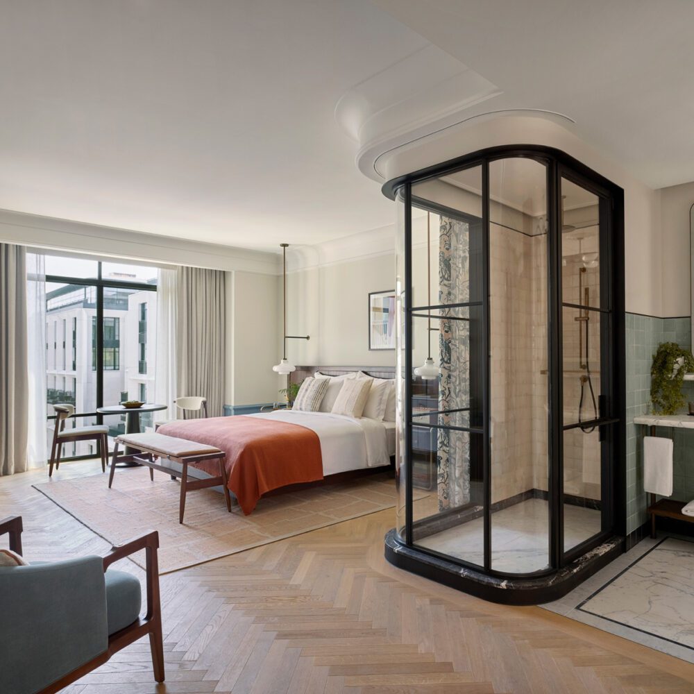 imgi_86_Premier___Premier_Terrace_Room by six senses london Spacious modern hotel room with a large bed, orange throw, glass enclosed shower, wooden armchairs, and floor-to-ceiling window with city view.