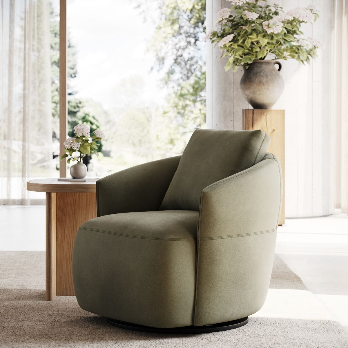 A cosy, olive-green armchair sits in a sunlit room next to a wooden table with flowers. Large windows reveal a serene, green outdoor view.