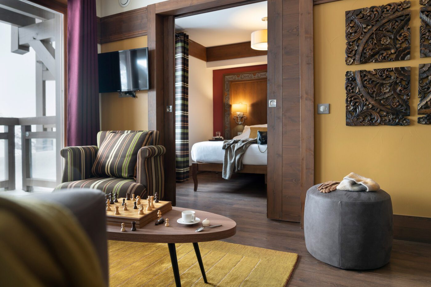 A modern hotel suite with a striped armchair, chessboard on a coffee table, tea set, and a view into a bedroom with a neatly made bed and decorative wall art.