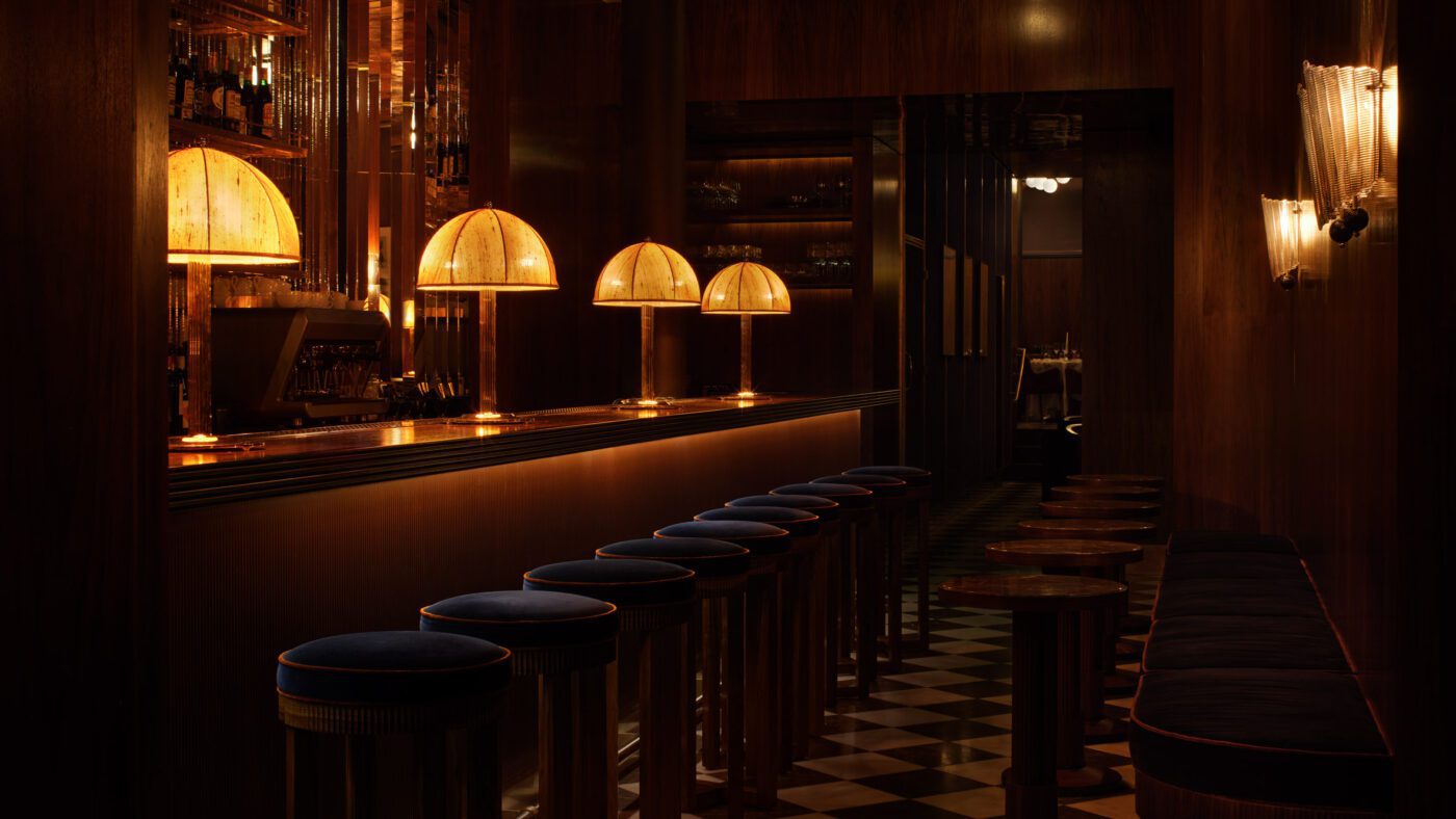 the-dover-restaurant-quincoces-drago-interiors-london-hero_dezeen_2364_col_0 A dimly lit bar with a warm, amber glow features dome-shaped lamps, dark wooden decor, plush bar stools, and checkered flooring, creating an intimate ambiance. One of the best designed restaurants in London