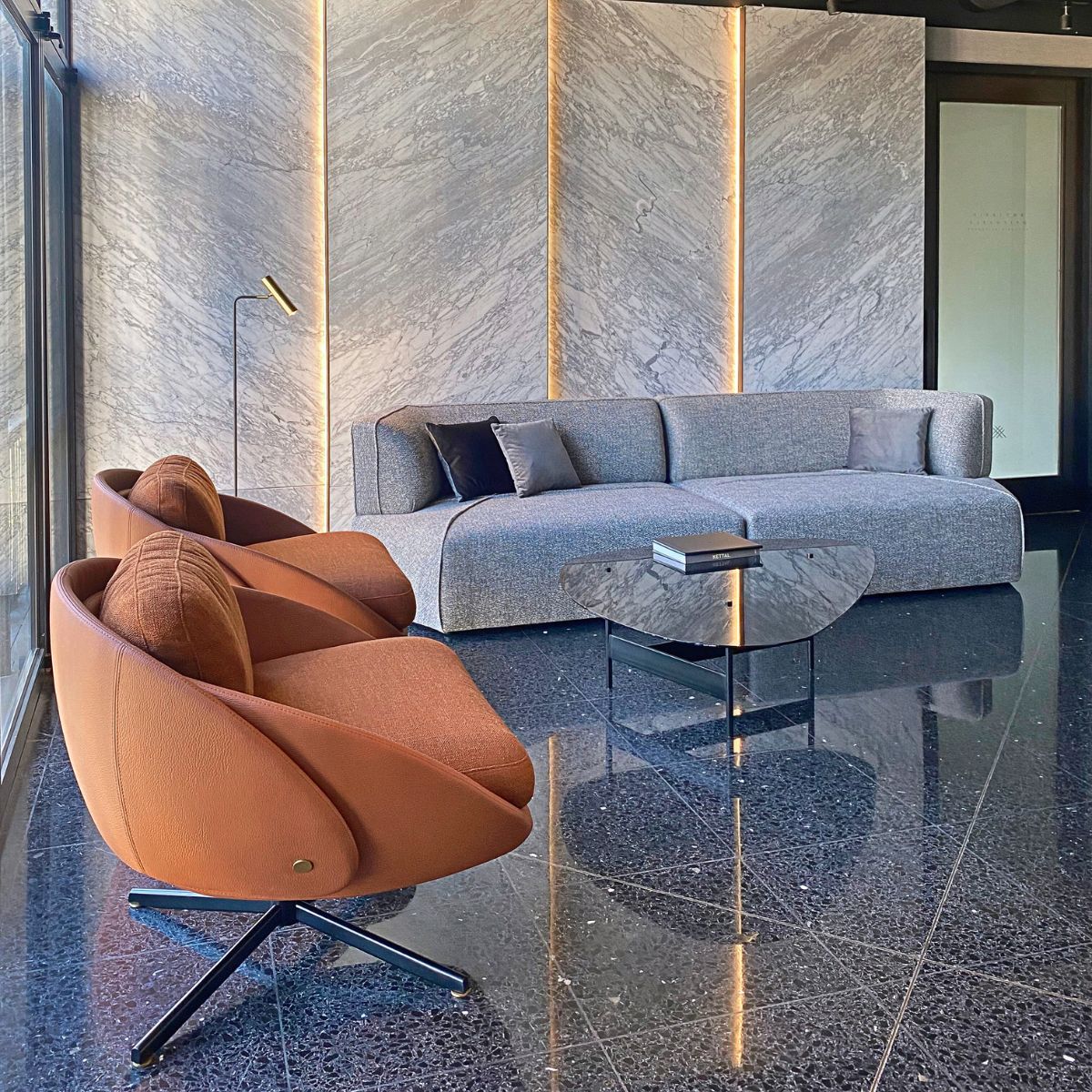 tres mas dos project Disruption Sofa (1) Modern living room with a gray sofa, two rust-colored swivel chairs, a sleek glass table, and marble wall panels with vertical lighting accents. By Tres Más Dos