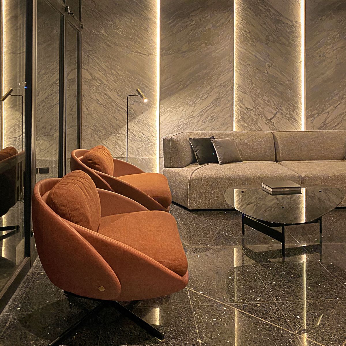 tres mas dos project Disruption Sofa Stylish modern office lounge with two orange chairs, a grey sofa, and a sleek glass table on a shiny black marble floor. Warm lighting creates a cosy atmosphere.
