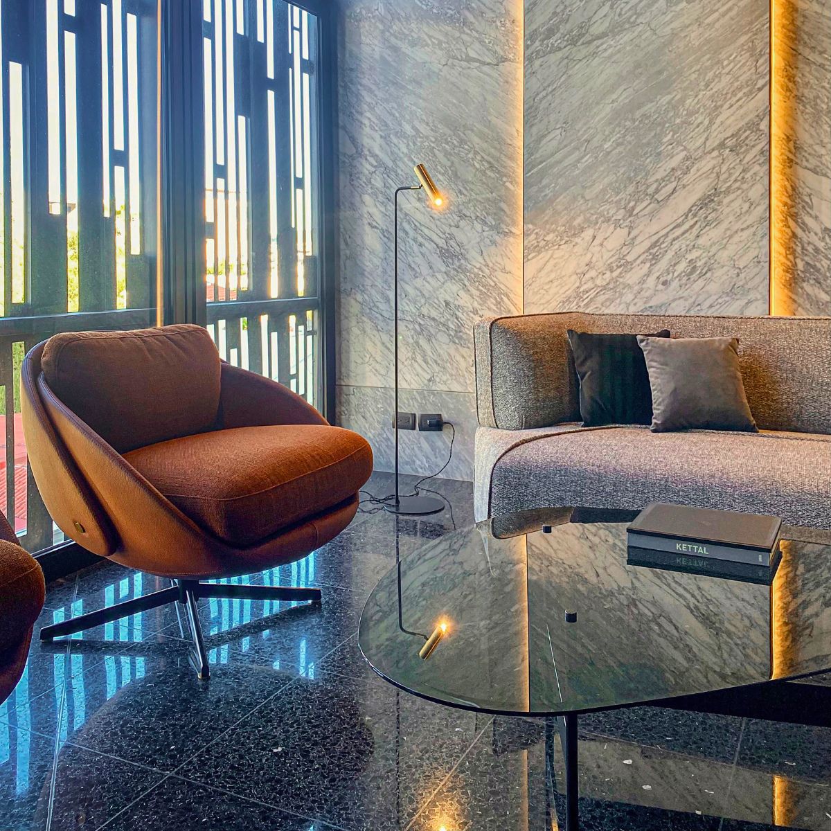 tres mas dos projetc Disruption Sofa (1) Stylish office lounge with a brown swivel chair, gray sofa, and modern floor lamp on marble floor. Sunlight streams through vertical window blinds. By Tres Más Dos