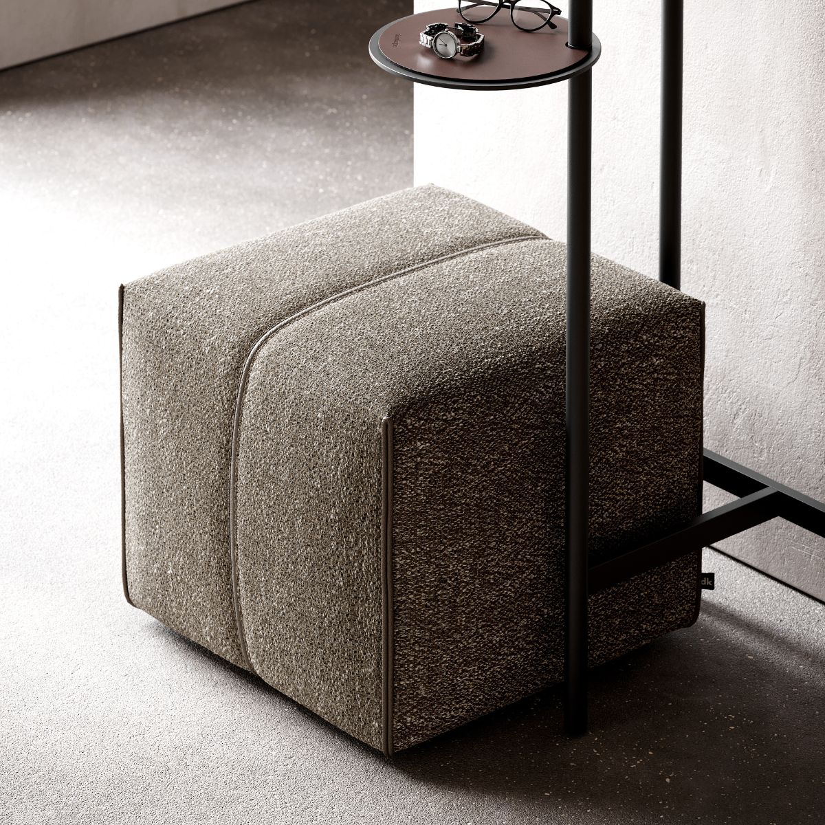 A textured gray pouf sits on a speckled floor next to a slim, black metal stand with a round shelf holding glasses and a watch. The setting feels modern and minimalist.