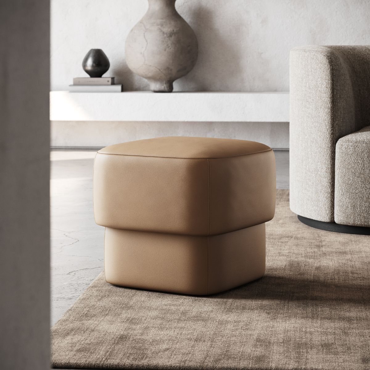 Modern interior with a minimalist, tan leather pouf on a beige rug. Nearby is a light gray sofa and a shelf with decorative vases, creating a calm ambiance.