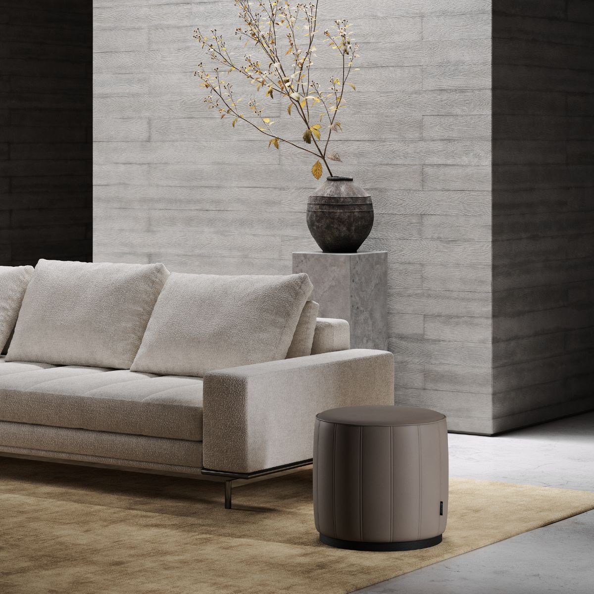 A modern living room with a beige sofa and a brown cylindrical pouf on a beige rug. A minimalist vase with branches sits on a concrete pedestal, creating a serene atmosphere.