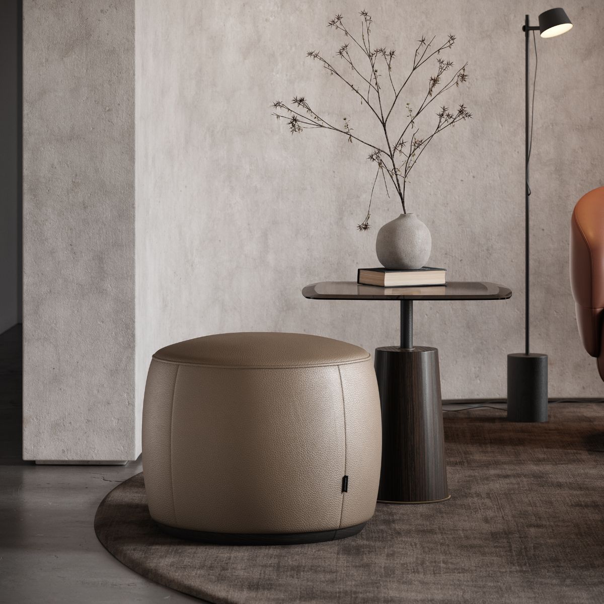 A modern living room with a beige pouf, a small round table holding a vase with twigs, a book, and a dim floor lamp, exudes a minimalist, calm vibe.