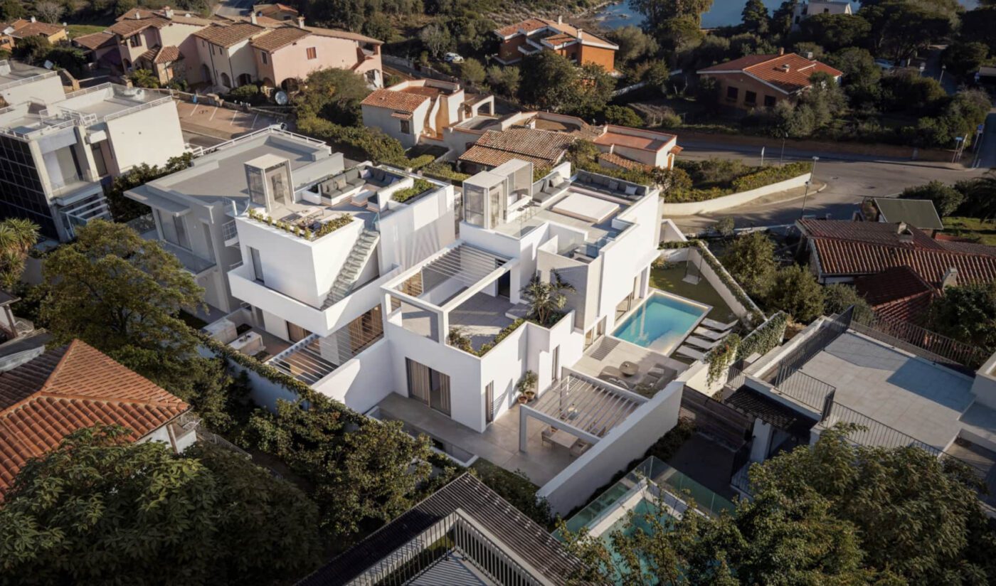 villa azulis aramis by rental 12 Aerial view of a modern, white cubic house with multiple terraces and a pool, surrounded by green trees and traditional homes in a suburban setting. By Rental 12.